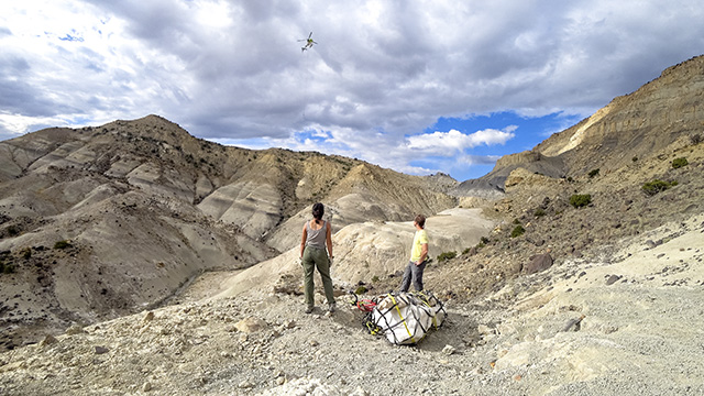 Dinosaur egg clutch in plaster resting on Utah desert with two paleontologists watching as helicopter arrives to airlift the fossils.