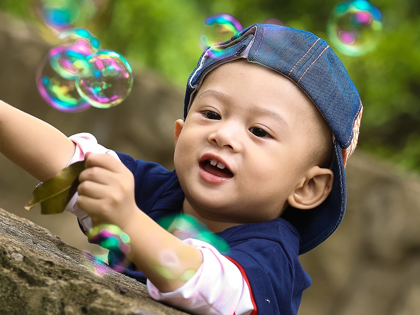 Baby in backwards hat plays with pretty pink and green bubbles outdoors.