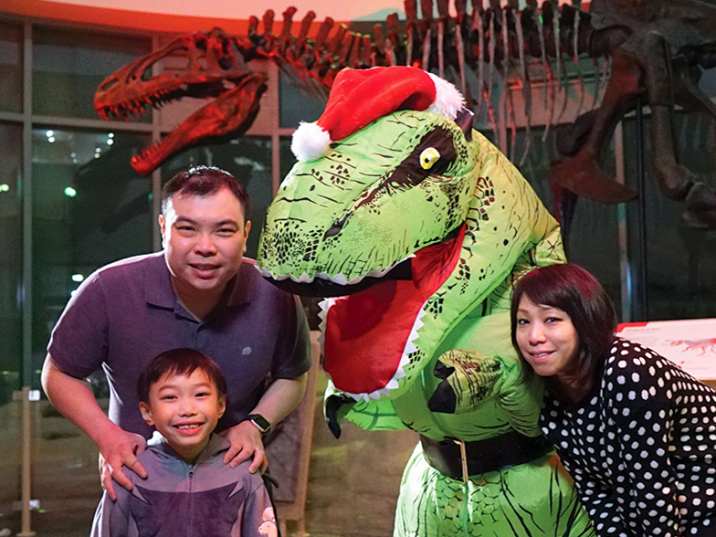 A family of 3 stands smiling around a dinosaur dressed in a red Santa Claus hat.