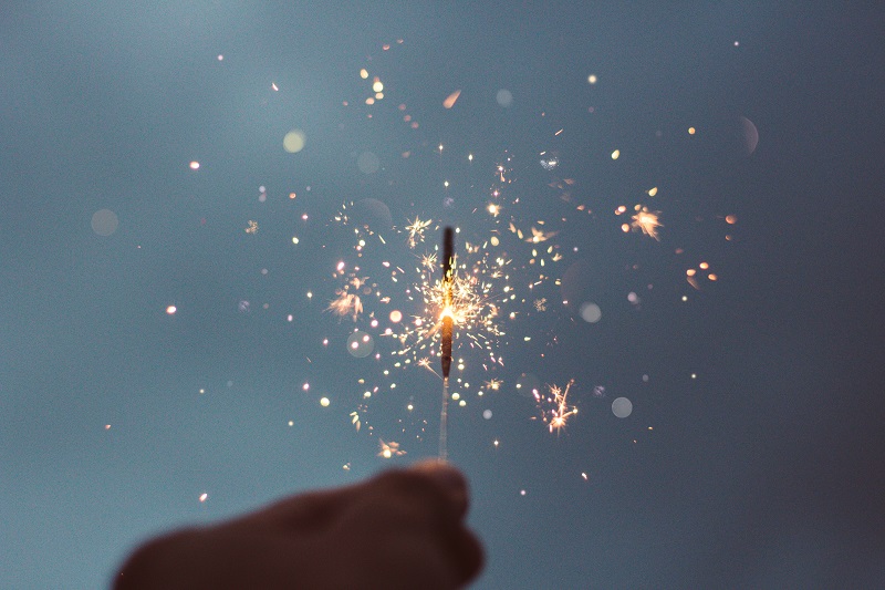 Sparklers glimmer different soft pastel colors from a sparkler. Pink, yellow, white and blue sparks shoot from the sparkler, held between the close-up fingers of a silhouette person. 