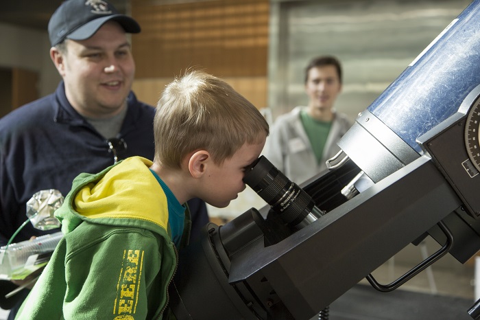 Young kid gazes into a microscope as his father smiles behind him. 