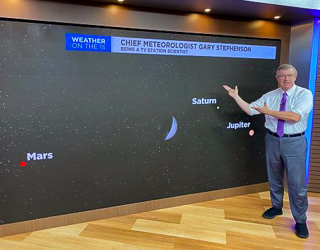 A meteorologist standing by a screen showing planets in the sky
