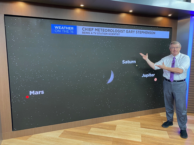 A meteorologist standing by a screen showing planets in the sky