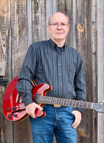 A white man leans against a weathered wooden wall. He is bald, wearing glasses, a blue, long-sleeved button up shirt and blue jeans. He is holding a brown electric guitar against his right hip. 