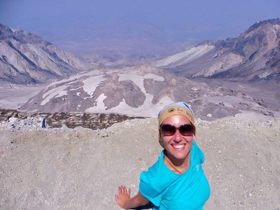 A woman in a blue shirt and bandana sits on a rocky ledge overlooking the volcano, Mount Saint Helens.