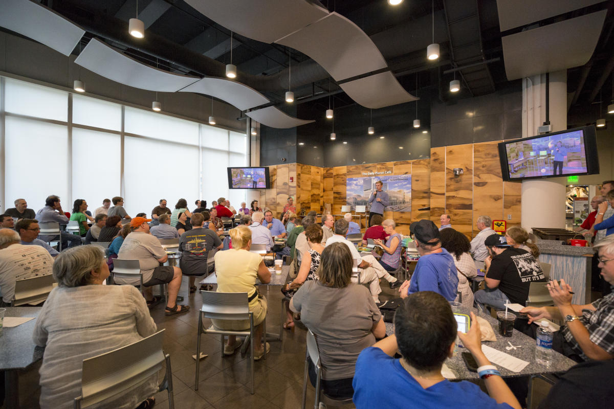 Photo of Cafe setting full of people sitting around tables with a person on a stage 