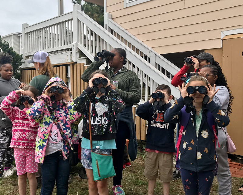 A group of adults and children looking for birds through binoculars in front of a house.