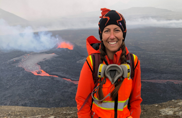 Photo of a woman wearing a bright orange coat and a safety vest standing high above a river of lava from a volcano