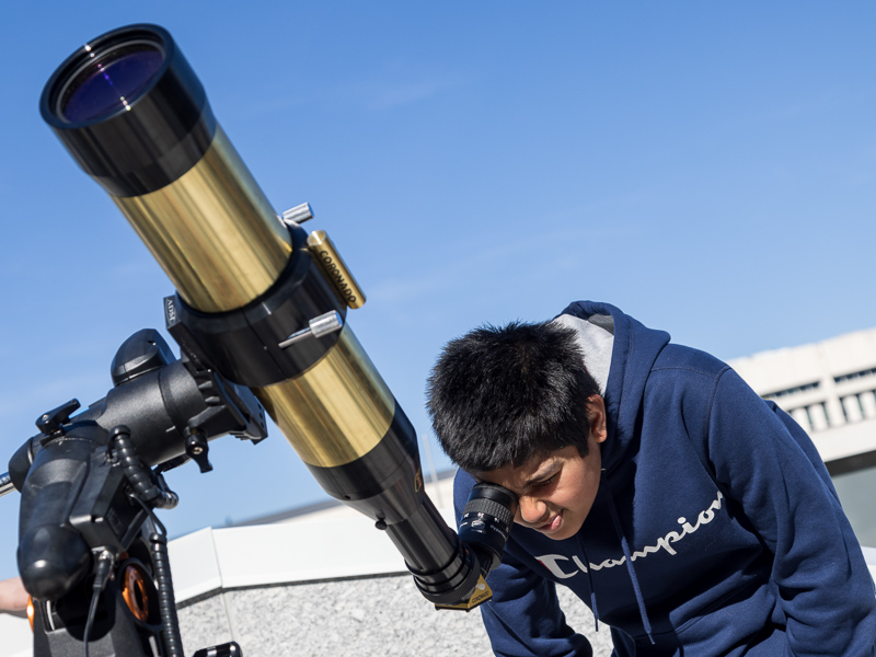Hispanic boy in a blue sweatshirt looks at the Sun through a special telescope.
