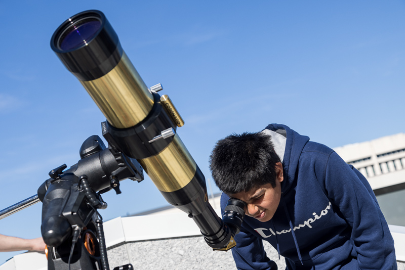 Light brown-skinned boy with black hair in a blue sweatshirt looks at the Sun through a special telescope.
