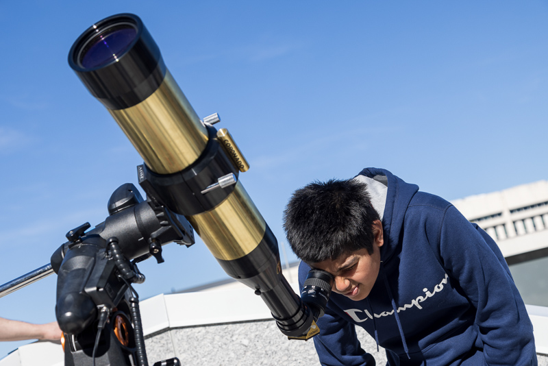 Hispanic boy in a blue sweatshirt looks at the Sun through a special telescope.