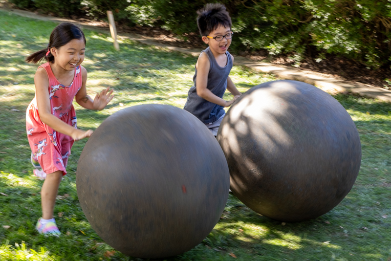 Two children race each other by rolling large 'dung balls' down a grass track.