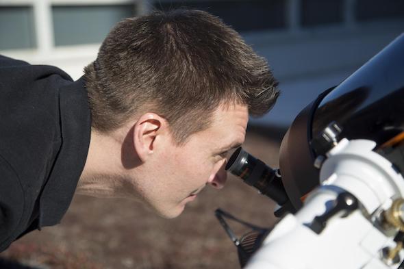 White male with short hair looks through the eye piece of a telescope.