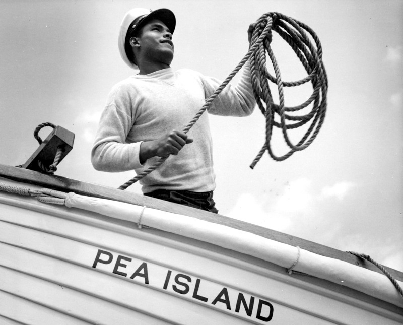 Black surfman standing with a coiled rope in a boat named Pea Island.