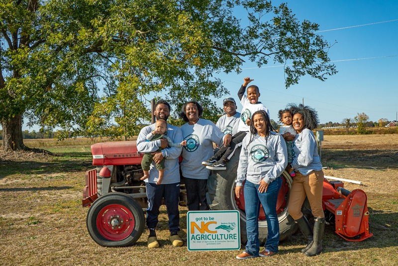 Members of the Black Family Land Trust standing in front of a tractor in a field, with a large tree on the left.