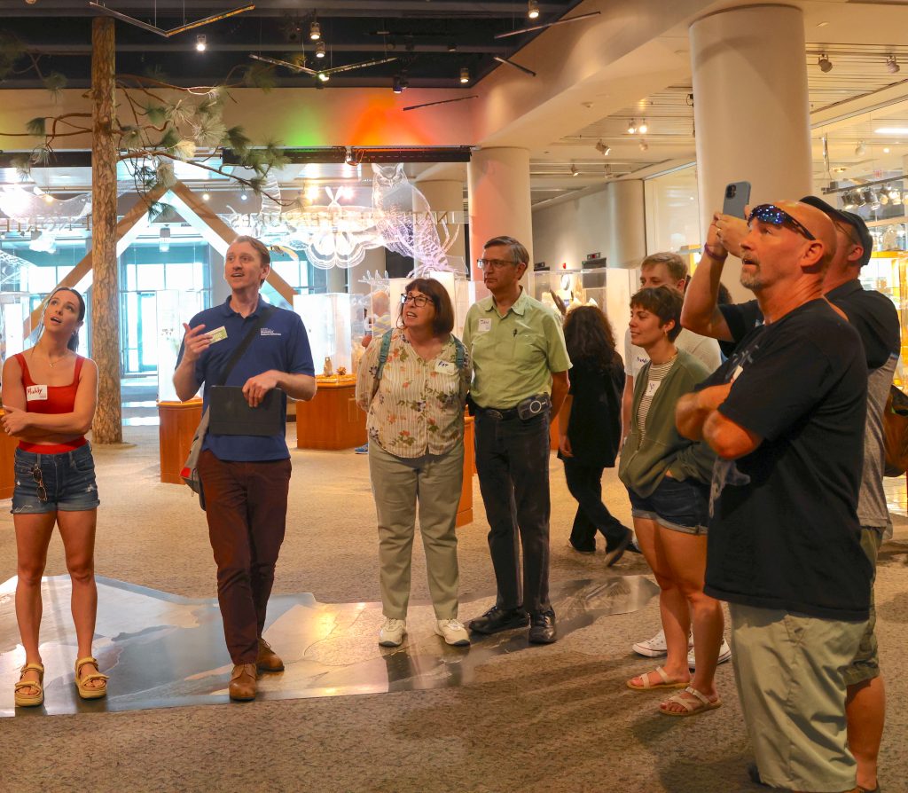 Image of a group of smiling adults look at an exhibit inside the Museum as a tour guide gestures