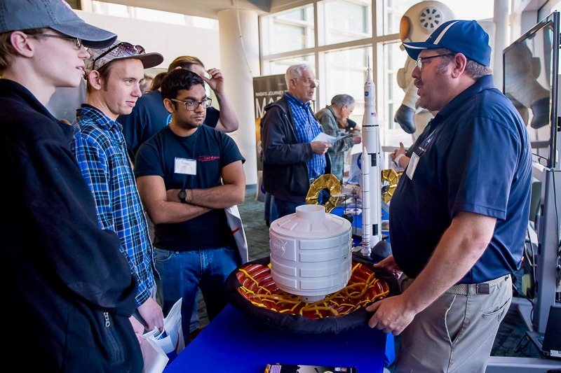 A NASA/JPL Solar System Ambassador speaks to a group of teens and adults over a white model of a spacecraft mechanical part.