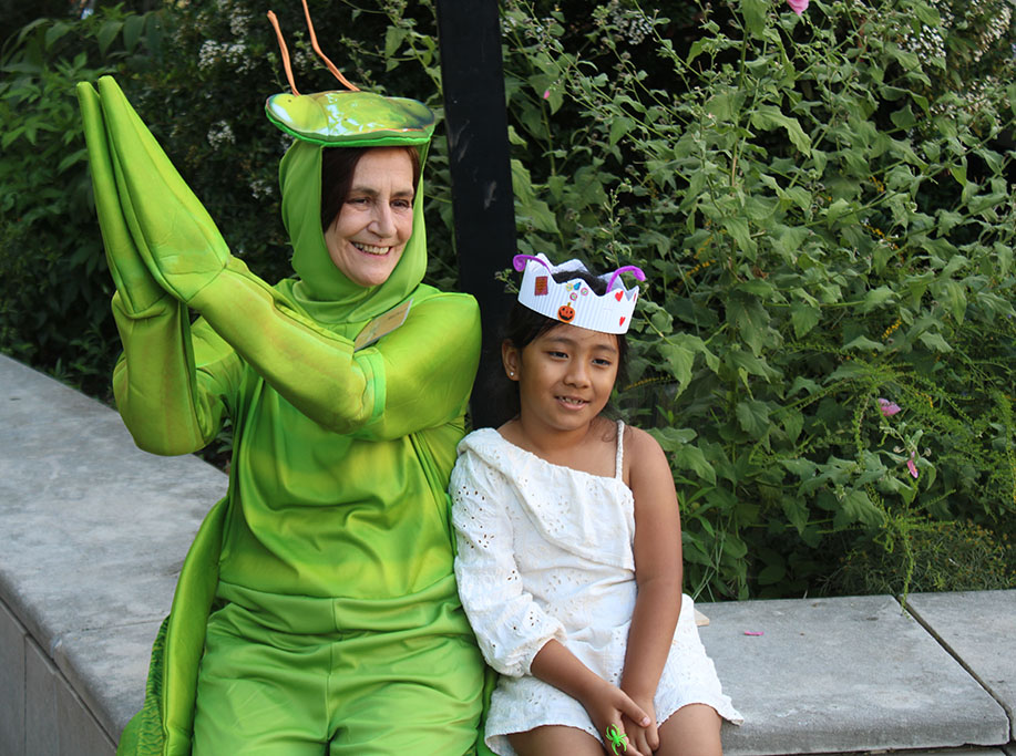 A woman dressed in a bright green grasshopper costume sits next to a young girl wearing a paper crown. 