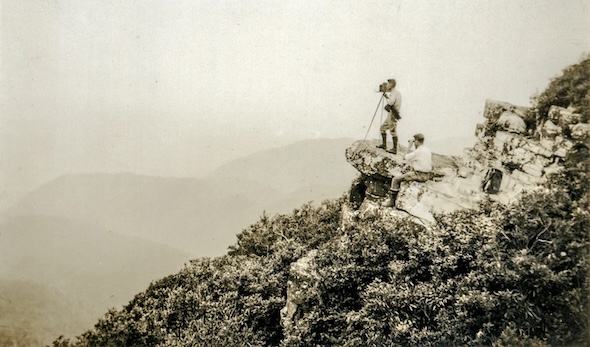 A black-and-white photograph of two men on a mountain side rock outcrop with an old camera on a tripod, with mountains in the distance