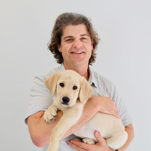 Brian Hare, a white male with brown hair, smiles for a portrait while holding a tan-colored puppy in his arms