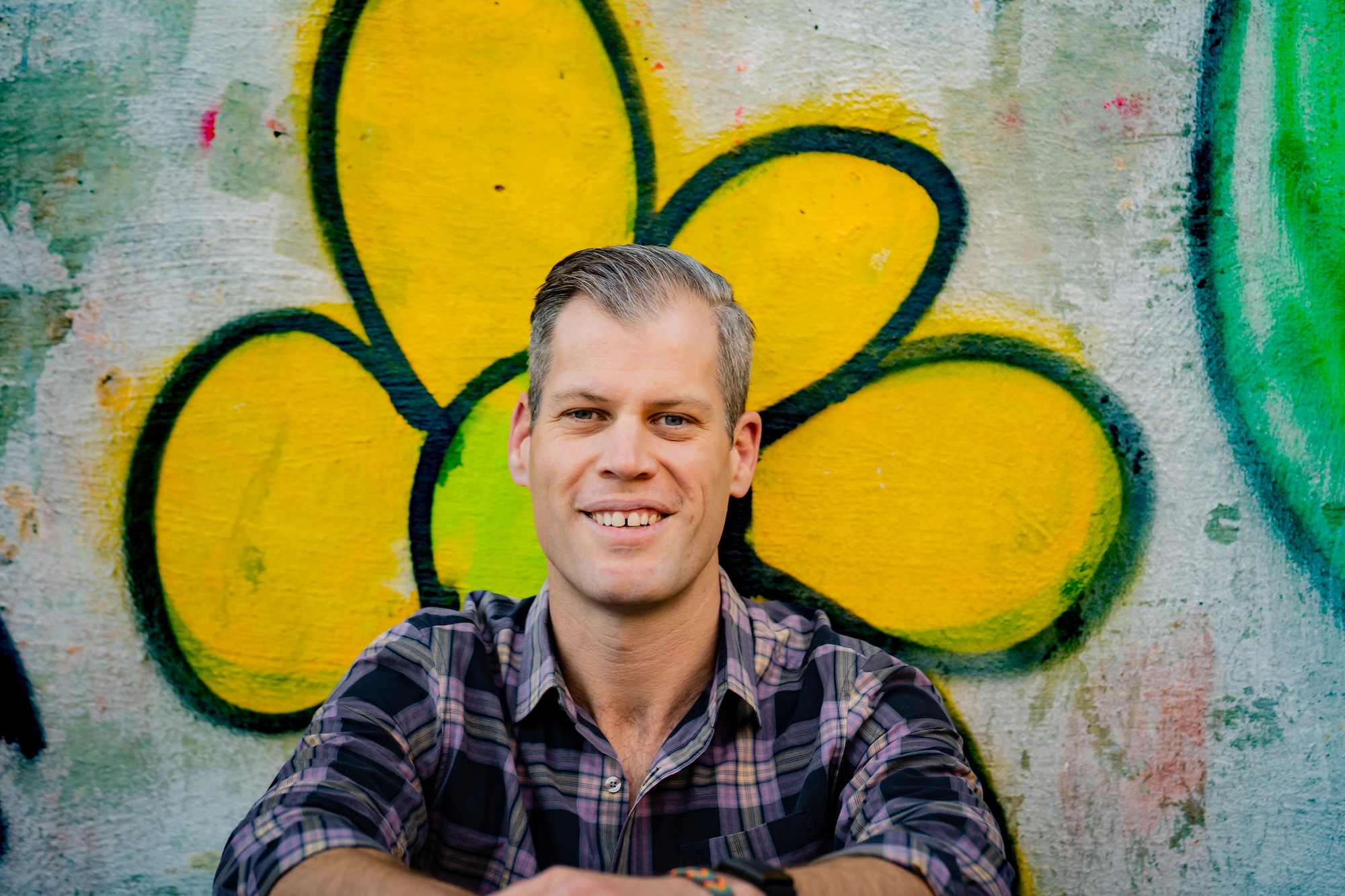 portrait of a man with light skin and combed back hair in a flannel shirt sitting in front of a graffiti flower 