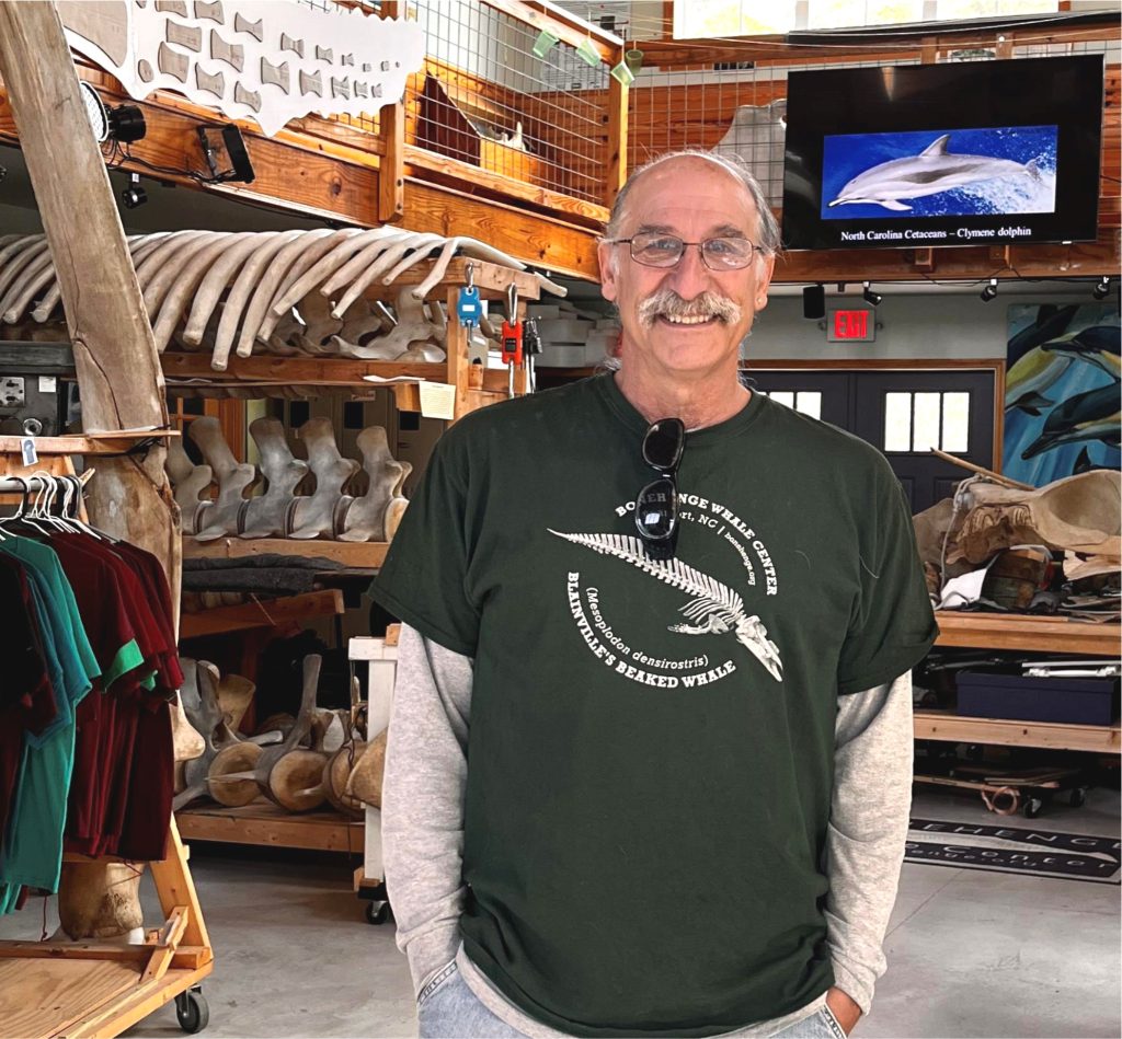 Close-up of Keith Rittmaster in smiling in a green shirt with a whale skeleton surrounded by whale skeletons in a workshop