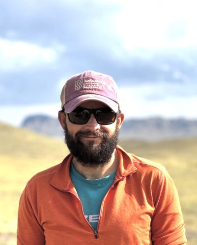 portrait photo of a bearded man wearing a pink hat that says Stellenbosch University, sunglasses and a green shirt with an orange jacket. Behind him is a mountainous desert landscape.