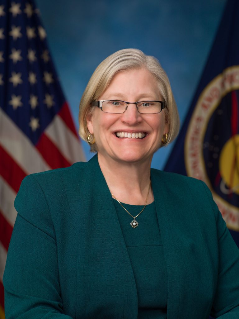 Photo of a woman with blond, chin length hair and glasses. Woman is smiling and wearing an emerald green shirt and suit jacket.