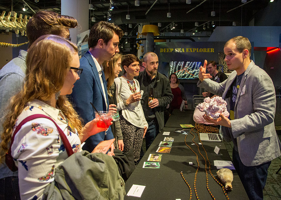 A group of young adults stand to the left of a table with a black tablecloth. Various lengths of rope and photographs are laying on the table. A man stands on the right side of the tables and is talking to the crowd. He is holding an anatomical model.