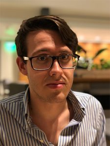 A mid-20s white man with medium length brown hair, black-framed glasses, and a blue-striped button up shirt. The background is blurred with the faint interior view with plants and lights.