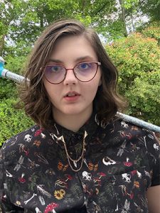 Young white woman with glasses and shoulder-length wavy brown hair, wearing a patterned button-up and holding a blue floral cane over their shoulder.