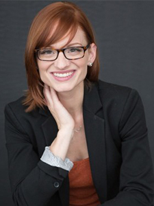 A photograph of Lacey, a redhead wearing glasses and a black blazer, with a small smile.