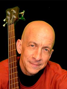 Ted smiling at the camera, wearing a red sweater over a black shirt and holding his bass guitar.
