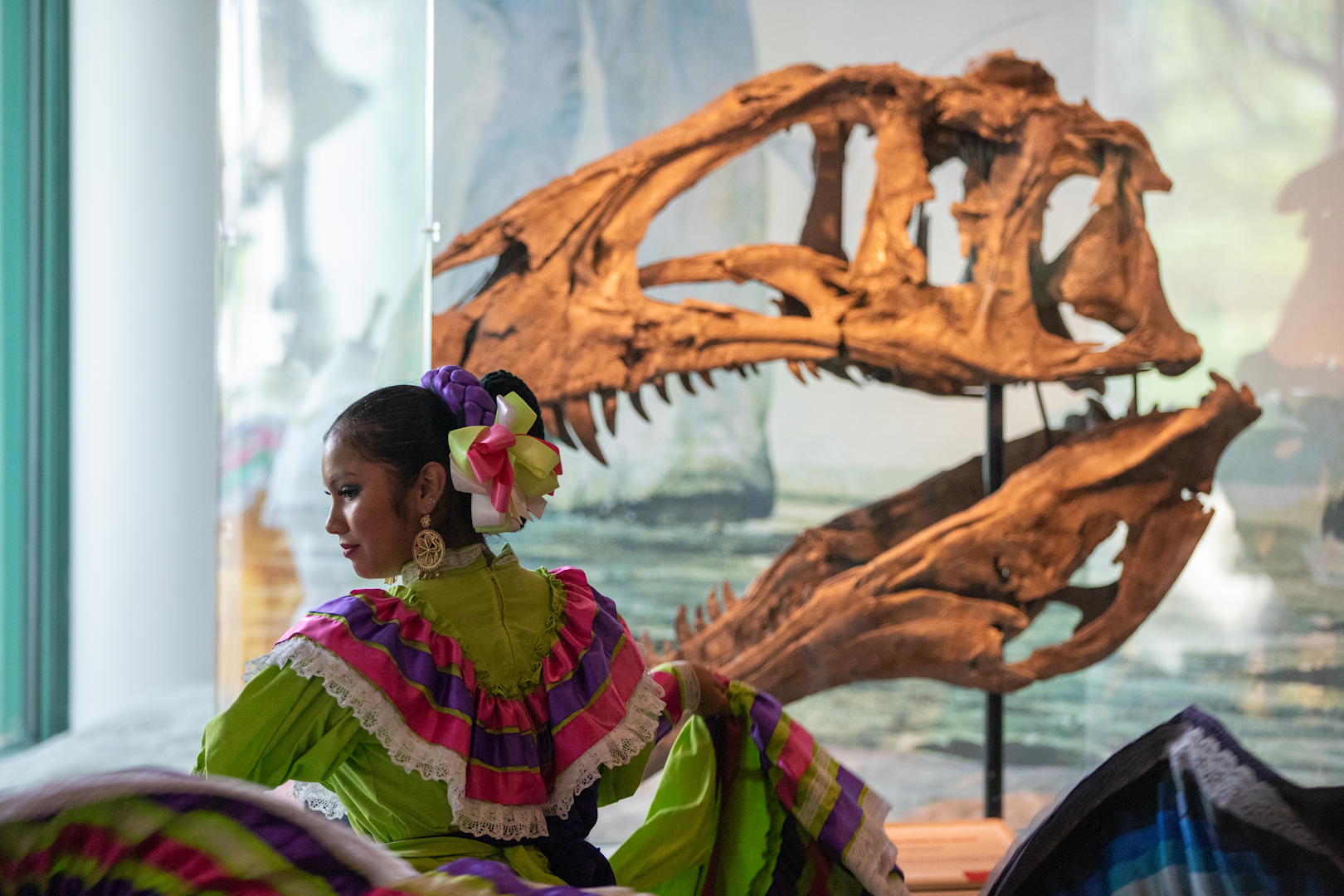 A woman in colorful garments dances with a model of a dinosaur skull in the background