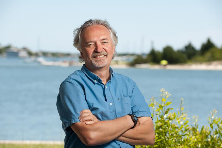 White male with white hair, wearing a blue shirt in front of a water background.