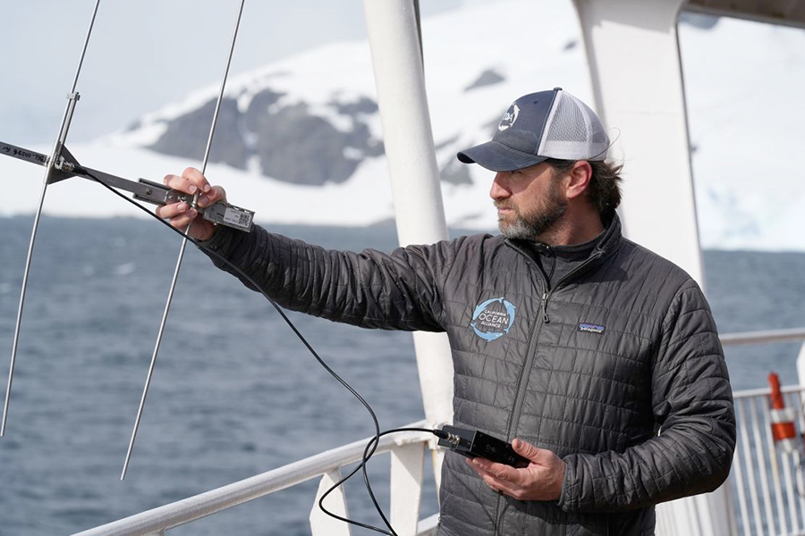 White man with brown hair and a beard wearing a grey coat and hat on a boat points an electronic sensor at the ocean with a snowy environment behind him.