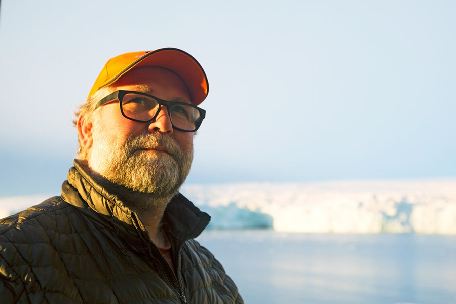 White man with a beard and dark-rimmed glasses, wearing an orange hat and green coat in front of a water environment.