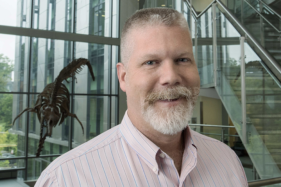 White man with a white beard standing in front of a fossil.