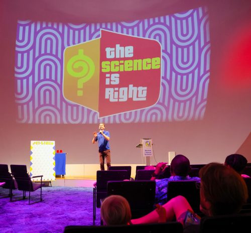 Chris Smith standing on a brightly lit stage with a graphic projected behind him that says The Science is Right in bright colors as a small audience watches.