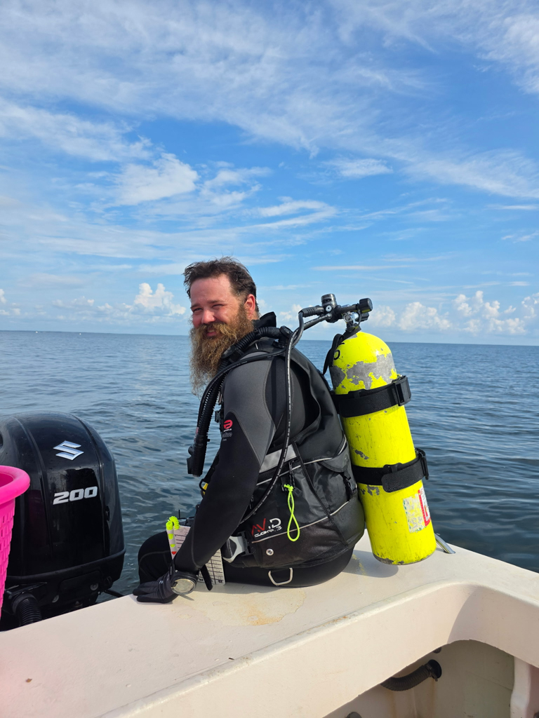 Man with a beard wearing a wetsuit and diving gear sitting on the edge of a boat