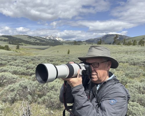 Mike Dunn holds a camera to his face with a sweeping landscape of grasses and mountains behind him