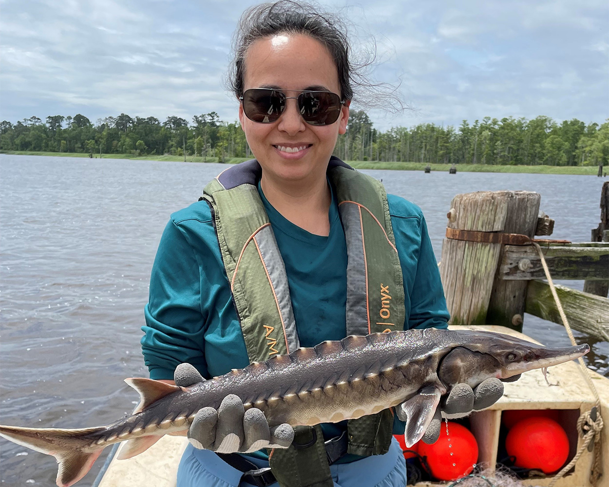 A woman on a dock holds a sturgeon, with a body of water in the background.
