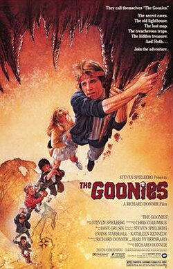 Movie poster for "The Goonies". The image is realistically-drawn and features 7 young teenagers holding onto each others legs. The first in the line is holding onto a stalactite.