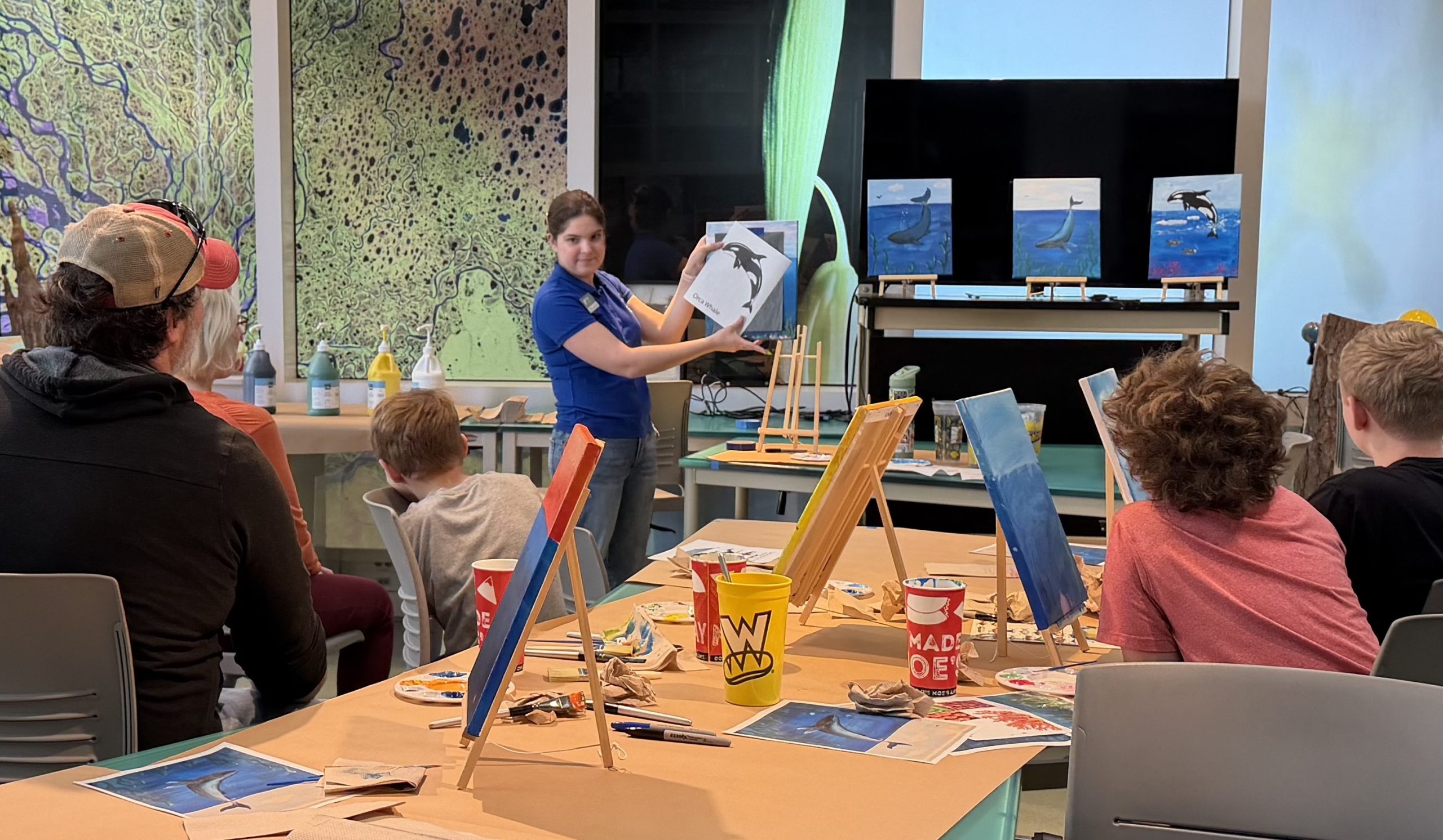 Three children and one adult man sit at a table with easels and painting supplies. A woman stands in the front of the room demonstrating the next step in a painting process.