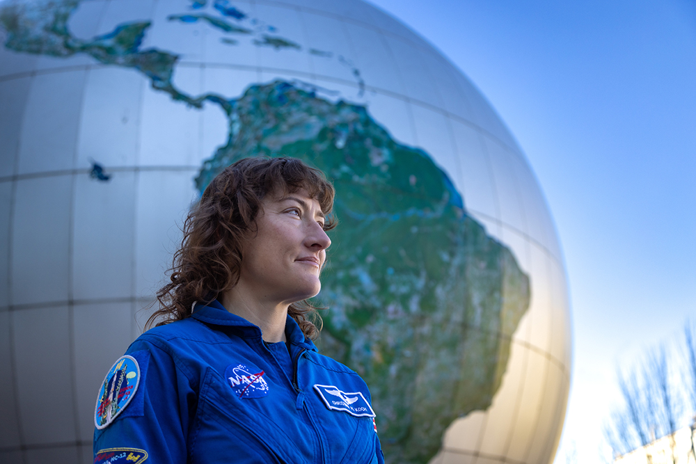 NASA Astronaut Christina Koch stands in front of the Daily Planet Theater, wearing a blue NASA suit.