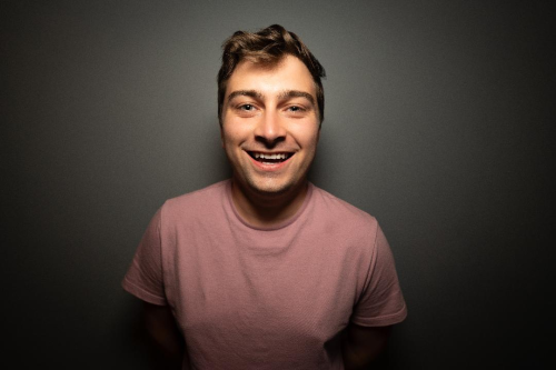 Portrait of Collin Brown smiling looking at the cmaera in a t-short against a dark gray background