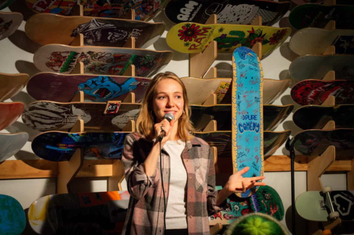 Stand-up comedian speaking into a microphone, gesturing with one hand, with a display of brightly illustrated skateboard decks behind her.