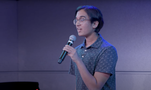 Photo of Nik Narain wearing glasses speaks into a handheld microphone against a blue-purple stage background.