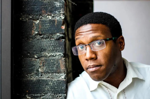 portrait of Will Thomas in glasses looking toward the camera next to a textured brick wall.